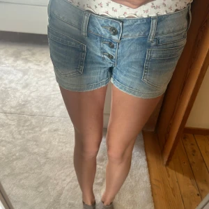 Trendiga lågmidjade Blå jeansshorts med knappar - Snygga blå jeansshorts med låg midja och flera framfickor samt bakfickor med knapp. Shortsen har en cool knappgylf och klassisk denim-look. Perfekta för varma dagar och enkel att matcha med olika toppar. W 28 dvs strl M
