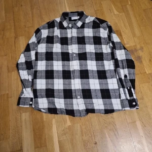 Svartvit rutig skjorta från ASOS DESIGN - Klassisk svartvit rutig skjorta från ASOS DESIGN i 100% bomull. Skjortan har långa ärmar, knäppning framtill och klassisk krage. Perfekt för en avslappnad och stilren look.