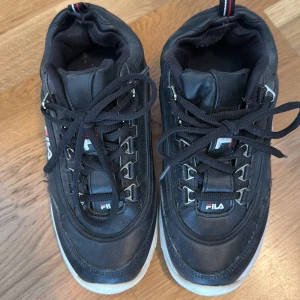 Svarta sneakers från Fila - Svarta Fila sneakers med vita sulor och svarta snören. Skorna har metallöglor för snörningen och Fila-logga på sidan, plösen och hälen. Klassisk sportig design med rund tå och vadderad kant för extra komfort.