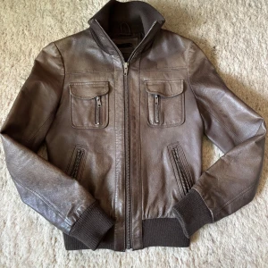 Brun skinnjacka med dragkedjor - Säljer en brun skinnjacka i äkta läder med ribbade muddar och krage. Jackan har fyra fickor framtill, alla med dragkedjor, och en klassisk bomberinspirerad passform. Perfekt för dig som vill ha en tidlös och cool look.