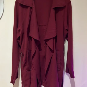 Vinröd kort kappa med bälte - Säljer min vinröda kappa med långa ärmar och ett matchande bälte i midjan. Jackan har en öppen front med draperad krage och två fickor framtill. Perfekt för lager-på-lager och enkel att styla till olika outfits.