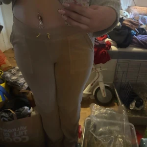 Beiga mjukisbyxor från Juicy Couture - Säljer ett par beiga mjukisbyxor från Juicy Couture i storlek L. Byxorna har dragsko i midjan med gulddetaljer och fickor bak med lock och knappar. Perfekta för en avslappnad och trendig look.