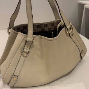 Beige handväska från Gucci - Säljer en snygg beige handväska från Gucci med klassiskt GG-mönstrat innerfoder. Väskan har två axelremmar, guldfärgade metalldetaljer och är tillverkad i skinn. Priset går att diskutera