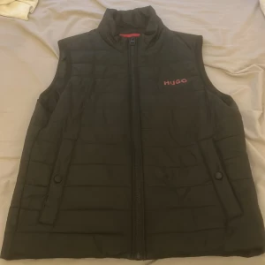 Svart dunväst från Hugo - Snygg svart dunväst från Hugo med diskret quiltad design och röd logotyp på bröstet. Västen har dragkedja framtill, ståkrage och två fickor med knappstängning. Perfekt för lager-på-lager och enkel att matcha. Västen kanske är använd 20 gånger Max eftersom den blev liten rätt så snabbt.