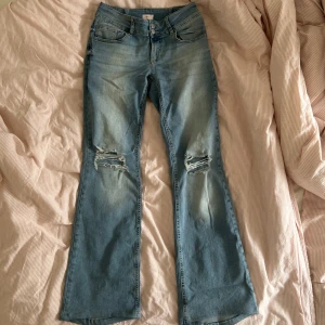 Ljusblå bootcut jeans med slitningar från Nelly - Säljer ett par ljusblå bootcut jeans från Nelly med slitna detaljer på båda knäna. Jeansen har klassisk femficksdesign och stängs med dragkedja och knapp. Perfekta för en avslappnad och trendig look. Jag har sytt in den i midjan lite.