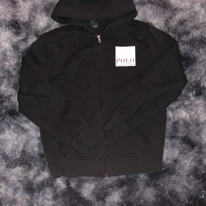 Svart hoodie från Polo Ralph Lauren - Svart hoodie med dragkedja från Polo Ralph Lauren. Stor vit logga på bröstet och ryggen. Klassisk huva och långa ärmar, tillverkad i mjuk bomull. Perfekt för en avslappnad stil.