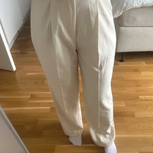 Beiga kostymbyxor med hög midja - Stilrena beiga kostymbyxor med hög midja och veck framtill. Byxorna har en rak passform och är perfekta för en dressad look. Tillverkade i ett mjukt material som ger en snygg siluett.
