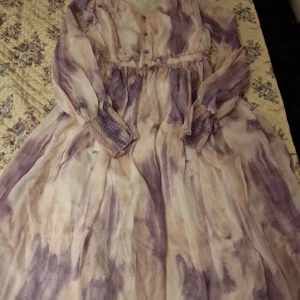 Långklänning i lila och beige mönster - Helt ny ej använd. Säljer en flowy långklänning med lila och beige mönster. Klänningen har långa ärmar med smock vid ärmsluten och knappar framtill. Materialet är tunt och luftigt, perfekt för varmare dagar. Snygg och trendig look med unikt mönster.