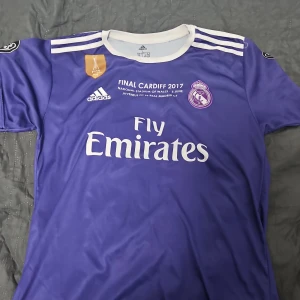 Real Madrid Champions League finaltröja 2017 Ronaldo - Lila Real Madrid fotbollströja från Champions League-finalen 2017 i Cardiff, med Ronaldo 7 på ryggen. Adidas-logga, klubbmärke och Fly Emirates-tryck på bröstet. Vita detaljer vid krage och ärmslut, samt final- och respektmärken på ärmarna.