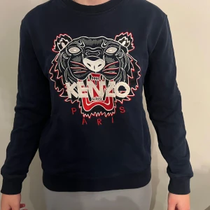 Mörkblå sweatshirt från Kenzo med tigertryck - Säljer en mörkblå sweatshirt från Kenzo med det ikoniska tigerhuvudet broderat framtill och texten 'Kenzo Paris' i vitt och rött. Tröjan har rund hals och långa ärmar. Perfekt statement-plagg för dig som gillar streetwear.