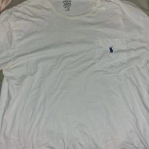 Vit t-shirt med bröstficka från Polo Ralph Lauren - Klassisk vit t-shirt från Polo Ralph Lauren med liten blå broderad logga på bröstfickan. T-shirten har rund hals och korta ärmar, perfekt för en clean och stilren look.