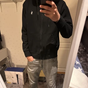 Svart hoodie från Polo Ralph Lauren - Svart hoodie från Polo Ralph Lauren med dragkedja och huva. Klassisk logga broderad på bröstet och två fickor framtill. Perfekt för en avslappnad stil.