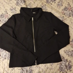 Svart ribbad tröja med dragkedja - Säljer en svart ribbad tröja med lång ärm och silverfärgad dragkedja framtill. Jackan har klassisk krage och är i ett stretchigt material som ger en snygg passform. Perfekt till många olika outfits! Helt ny med tagg.