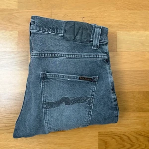 Grå jeans från Nudie Jeans Lean Dean - Säljer ett par grå jeans från Nudie Jeans med klassisk femficksdesign och svarta detaljer på bakfickorna. Jeansen har normal passform och raka ben, perfekt för en avslappnad stil.