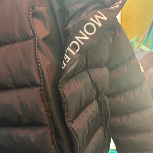 Svart dunjacka från Moncler - Säljer en svart dunjacka från Moncler med tydlig logotyp på ärmen och dragkedja framtill. Jackan har en vadderad, pufferstil och hög krage med Moncler-tryck. Perfekt för kyliga dagar.