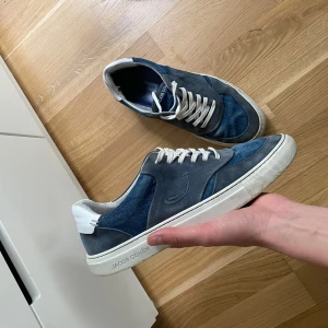 Blå sneakers från Jacob Cohën - Säljer ett par snygga blå sneakers från Jacob Cohën med vita snören och vit sula. Skorna har en mix av mocka och textilmaterial, samt diskret logga på sidan. Perfekta för en avslappnad och stilren look.