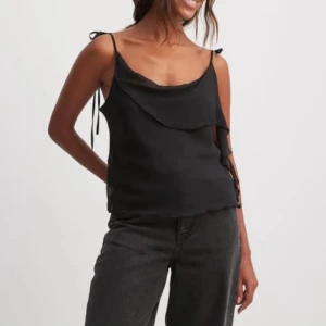 Volangtopp - Säljer en svart linnetopp med tunna axelband som knyts på axlarna och en snygg volangdetalj framtill och baktill. Toppen har en lös passform och är perfekt för varma dagar eller fest