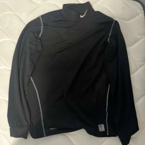 Svart långärmad träningströja från Nike - Svart långärmad träningströja från Nike med gråa detaljer och hög krage. Perfekt för träning eller löpning under kallare dagar. Tillverkad i syntetmaterial för bra andningsförmåga.