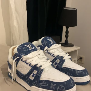 Louis Vuitton blå och vita sneakers - Säljer ett par Louis Vuitton sneakers i blått och vitt med tydlig LV-logga och monogrammönster. Skorna har vita snören, rund tå och är tillverkade i en mix av denim och skinn. Perfekta för dig som vill sticka ut med exklusiva detaljer.