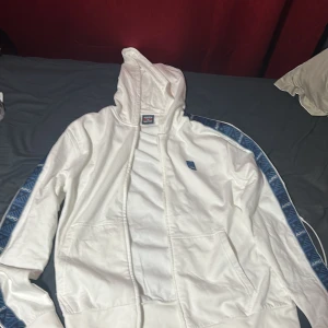 Vit hoodie från Aspen Polo Club med blå detaljer - Säljer en vit hoodie från Aspen Polo Club med blå logotyp på bröstet och blå mönstrade band längs ärmarna. Jackan har huva och dragkedja framtill, samt två fickor. Perfekt för en sportig och avslappnad stil.
