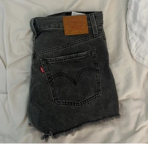 Svarta jeansshorts från Levi's - Snygga svarta jeansshorts från Levi's med slitna detaljer och fransig kant. De har en klassisk femficksdesign och knappar i gylfen. Perfekta för en avslappnad stil. Står tyvärt inte storlek högmidjade, första bilden är lånad