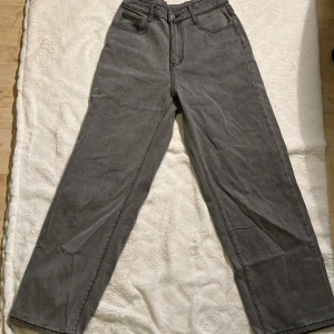 Gråa raka jeans med hög midja - Säljer ett par gråa jeans med raka ben och hög midja. Byxorna har klassiska fem fickor och synliga sömmar. Perfekta för dig som gillar en avslappnad och stilren look.