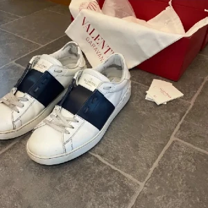 Valentino Garavani Open - Säljer ett par vita Valentino Garavani Open sneakers med bred mörkblå rem över mitten. Skorna har rund tå, platt sula och klassisk snörning. Perfekta för dig som gillar stilrena och exklusiva sneakers. 