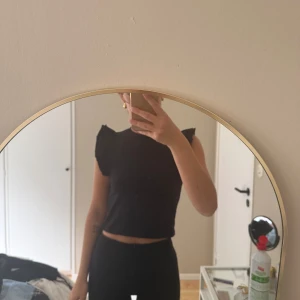 Svart croppad topp med volangärmar - Säljer en svart croppad topp med volangdetaljer på axlarna. Toppen är ärmlös och har en tight passform, perfekt att styla med höga byxor eller kjol.