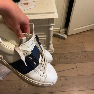 Valentino Garavani Open sneakers vit/blå - Snygga Valentino Garavani Open sneakers i vitt skinn med bred marinblå rem över mitten. Klassisk rund tå, platt sula och coola nitar baktill på hälen. Skorna har vita snören och diskret logga på plösen. Perfekta för dig som gillar exklusiva och stilrena sneakers.