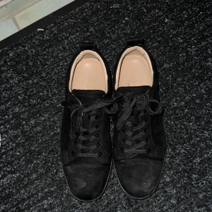 Svarta Christian Louboutin - Snygga svarta sneakers från Christian Louboutin i mocka med klassisk röd sula och guldfärgad logga på innersulan. Skorna har rund tå, svarta skosnören och en stilren look som sticker ut med den ikoniska röda undersidan.