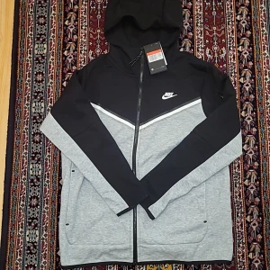 Nike svart och grå vindjacka L - Nike vindjacka i svart och ljusgrått med huva och hel dragkedja. Snygg V-detalj på bröstet, två fickor och klassisk Nike-logga på vänster sida. Jackan är i sportig stil och passar perfekt till streetwear. Materialet är troligtvis polyester.