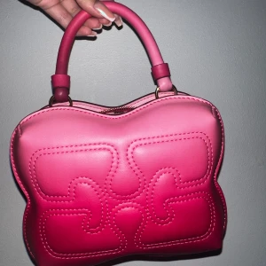 Rosa handväska från Ganni - Ganni Gradient Small Butterfly Crossbody Bag i Pink. Väskan har både handtag och justerbar axelrem och kommer tillsammans med en dust bag. Perfekt för dig som vill sticka ut med en färgglad accessoar.