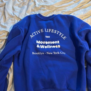 Blå sweatshirt Movement & Wellness - Blå sweatshirt med vit text på både bröstet och ryggen. Texten lyder 'Active Lifestyle 1990 Movement & Wellness Brooklyn - New York City'. Tröjan har rund hals, långa ärmar och ribbade muddar. Perfekt för en chill och sportig look.