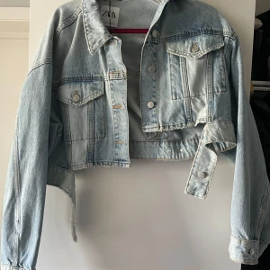 Croppad jeansjacka från Zara - Snygg croppad jeansjacka från Zara i ljusblå denim. Jackan har klassiska bröstfickor med knappar, krage och långa ärmar. Perfekt för dig som vill ha en trendig och avslappnad look.