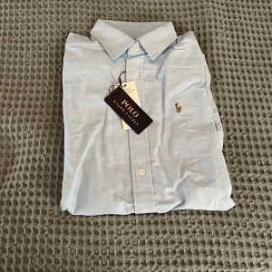 Ljusblå skjorta från Polo Ralph Lauren - Klassisk ljusblå skjorta från Polo Ralph Lauren med broderad logga på bröstet. Skjortan har knappar framtill och lång ärm med manschett. Perfekt för en stilren look. Öppen för förhandling.