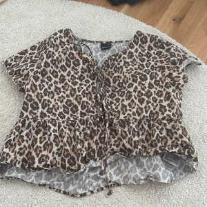 Leopardmönstrad blus från Gina Tricot - Säljer en kortärmad blus från Gina Tricot med leopardmönster i bruna och svarta toner. Blusen har volang nertill och knytning framtill för en snygg detalj. Perfekt för dig som gillar djurmönstrat och vill sticka ut.