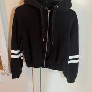Svart hoodie med vita ränder från H&M - Säljer en svart hoodie från H&M med dragkedja och två vita ränder på varje ärm. Tröjan har huva med snören och är croppad i modellen. Perfekt för en avslappnad stil.