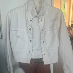 Vit jeansjacka  - Säljer en vit croppad jeansjacka från H&M med croppad passform och silverfärgade knappar. Jackan har två bröstfickor och slitna detaljer på framsidan. Perfekt för att lyfta en enkel outfit.