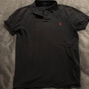 Svart pikétröja Polo Ralph Lauren  - Svart pikétröja från Polo Ralph Lauren i slim fit med klassisk krage och korta ärmar. Tröjan har det ikoniska röda broderade logotypen på bröstet och är tillverkad i bomull. 