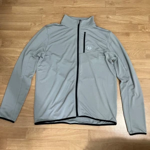 Ljusgrå vindjacka med dragkedja - Ljusgrå vindjacka med svart hel dragkedja och hög krage. Jackan har en diskret logga på bröstet och en bröstficka med dragkedja. Tillverkad i lätt syntetmaterial, perfekt för blåsiga dagar. Långärmad och med enkel, clean design. Den är från lager 157.