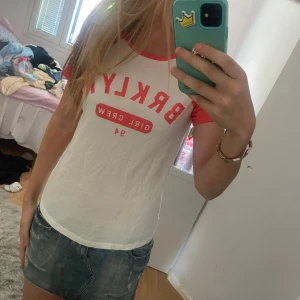 Vit och röd t-shirt från H&M med tryck - Säljer en vit t-shirt från H&M med coral ärmar och krage. Tryck på bröstet med texten 'BRKLYN GIRL CREW 94' i rött. Klassisk passform och kortärmad, perfekt till sommaren.