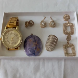 Mix av smycken och klocka i guld och lila - Säljer endast som set , smycken: ett stort guldigt armbandsur(nya behövs batteriet, ett lila hänge med blommotiv( handmade) med signature, ett par glittriga örhängen, ett par runda örhängen, ett stort glittrigt ring och ett par stora rektangulära örhängen med stenar.