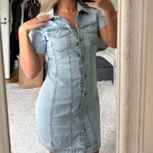 Ljusblå jeansklänning från Asos - Snygg ljusblå jeansklänning med kort ärm och markerad midja. Klänningen har klassisk skjortkrage, två bröstfickor med knappar och hel knäppning framtill. Den är figurnära och slutar ovanför knäna.