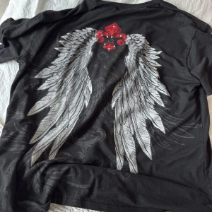 Svart t-shirt med vingar och röd detalj - Cool svart t-shirt med stort tryck av vita änglavingar på ryggen och en röd dekorativ detalj upptill. Klassisk passform och korta ärmar, perfekt för dig som gillar edgy och unika prints. Mjuk och skön bomullskänsla.