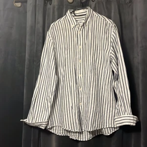 Linneskjorta från ZARA - Snygg vit skjorta från Zara med mörkblå vertikala ränder och relaxed fit. Skjortan har klassisk krage, långa ärmar och knäppning framtill. Tillverkad i linne perfekt för en chill och stilren look inför sommarn! Endast använd ett fåtal gånger så mycket bra skick.