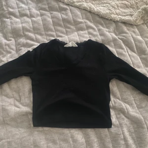Svart ribbad topp med spets från H&M - Svart långärmad ribbad topp från H&M med v-ringning och fina spetsdetaljer längs halsringningen. Croppad modell som sitter snyggt och passar perfekt till jeans eller kjol. Enkel men ändå med en extra touch tack vare spetsen.