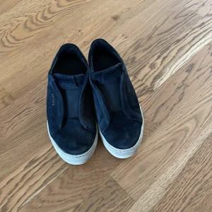 Svarta sneakers från Axel Arigato - Snygga svarta sneakers från Axel Arigato i mocka med vit platt sula. Minimalistisk design utan snörning och diskret logga i guld på sidan. Perfekta för en clean och stilren look. Ett litet hål har dem men annars är dem i bra skick. Vi kan diskutera pris