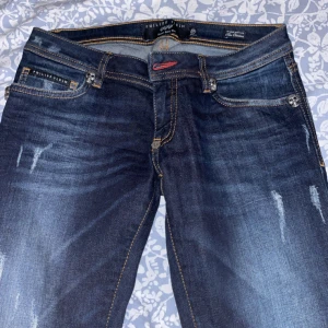Philipp Plein Super Sexy Slim jeans - Mörkblå slim fit jeans från Philipp Plein med snygga slitningar, orange kontrastsömmar och coola dödskalle-detaljer i metall vid fickorna. Modellen heter Super Sexy Slim och har en klassisk femficksdesign samt metallbricka bak.