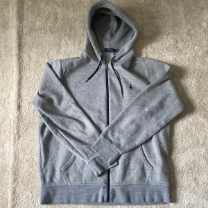 Grå hoodie från Polo Ralph Lauren - Säljer en klassisk grå hoodie från Polo Ralph Lauren med dragkedja och huva. Tröjan har svarta snören, två fickor framtill och den ikoniska broderade loggan på bröstet. Perfekt för en avslappnad stil.