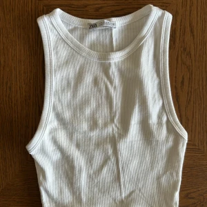 Vit ribbad linnetopp från Zara - Säljer en klassisk vit ribbad linnetopp från Zara. Toppen har rund halsringning och är ärmlös, perfekt för varma dagar eller som lager under andra plagg. Enkel och stilren design som passar till mycket. Använd en gång 
